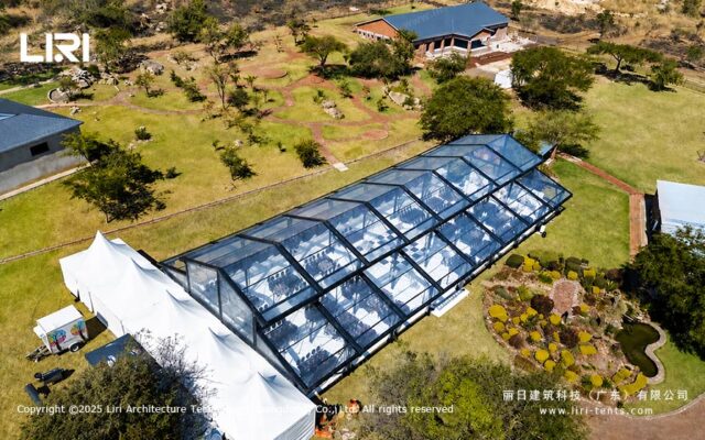 Vista aérea de uma tenda tipo orangerie para eventos ao ar livre, com telhado transparente e cenário de jardim